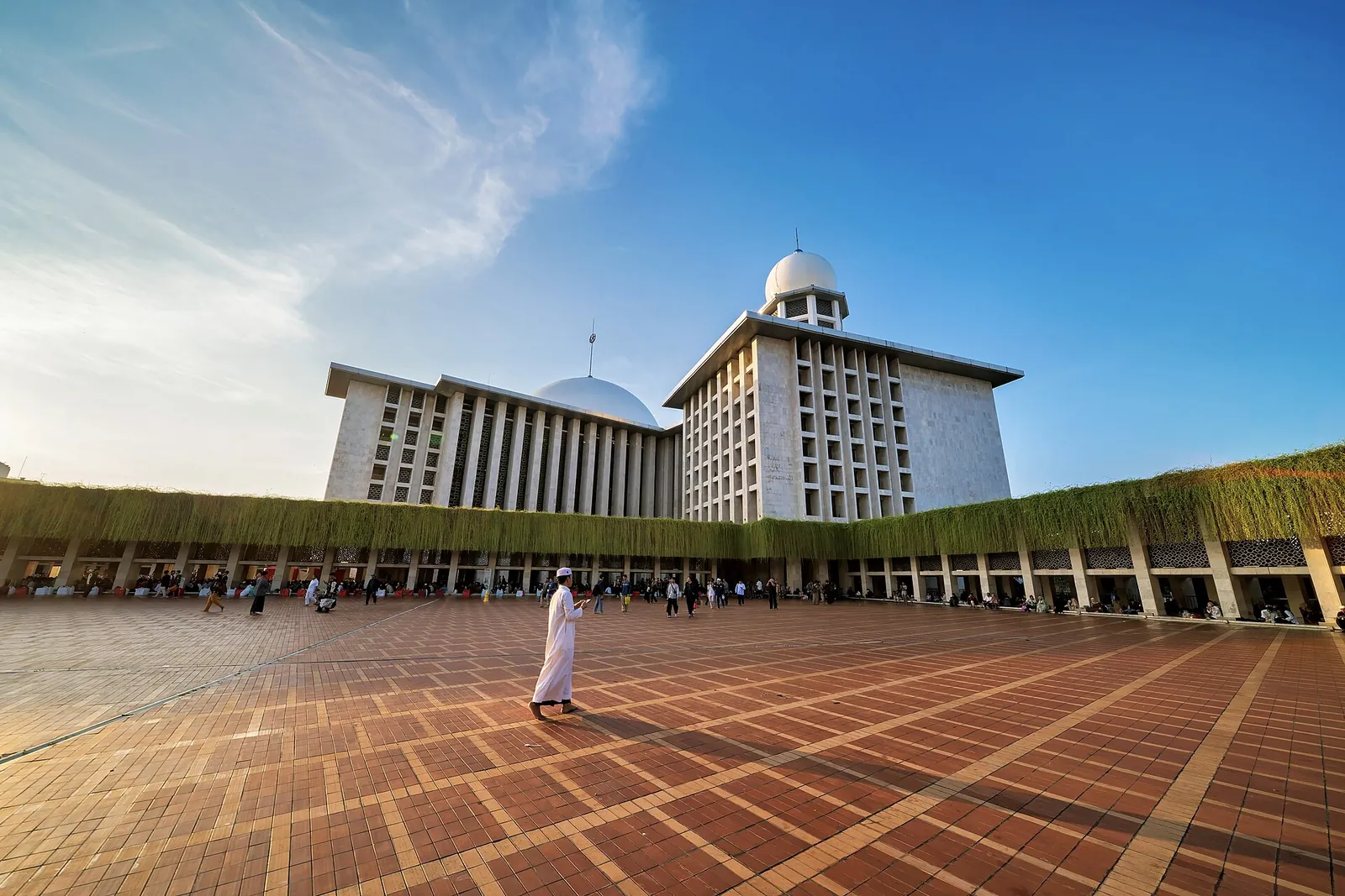 The white main hall and minaret of Istiqlal Mosque in Jakarta under a blue sky, with a wide paved courtyard