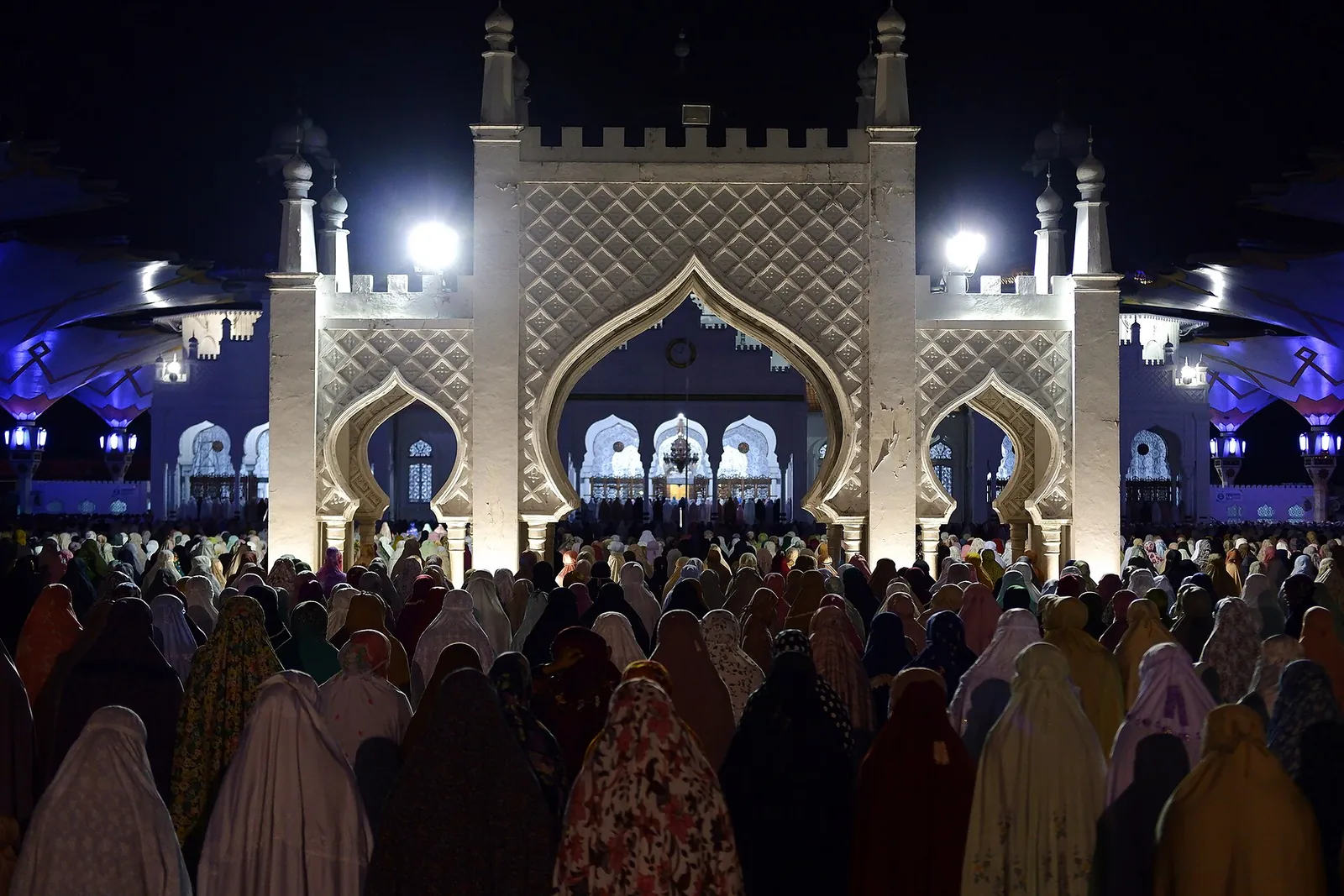 A large crowd of worshippers in white prayer robes before the floodlit gateway of Baiturrahman Grand Mosque at night