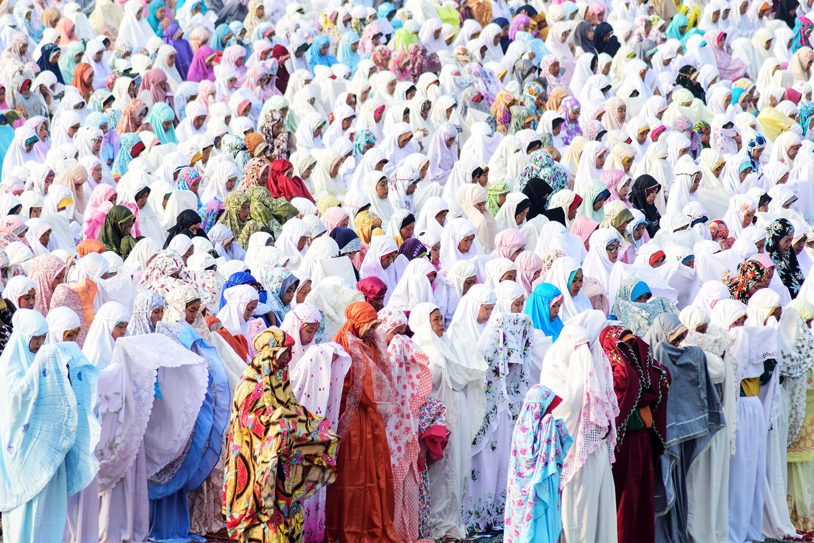 Rows of women in white prayer robes and men praying together on an open sandy beach
