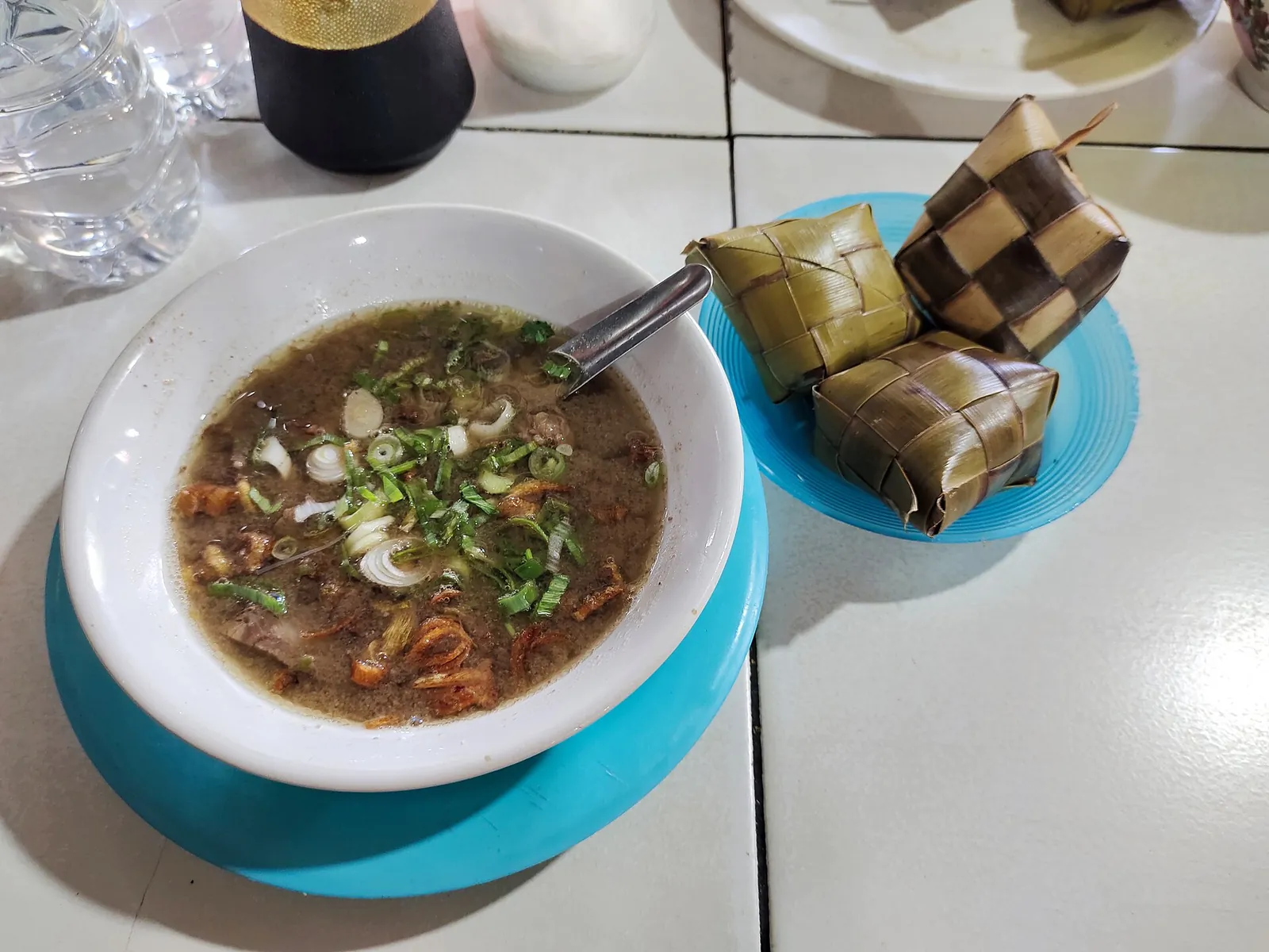A bowl of coto Makassar with spring onions next to woven ketupat rice parcels