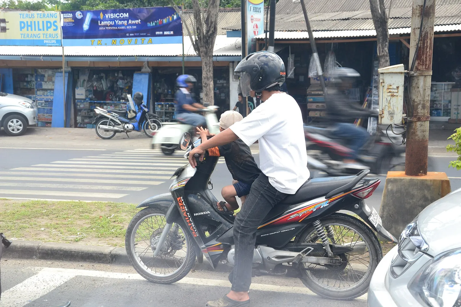 A man riding a motorbike in Bali traffic with a child sitting in front of him without a helmet