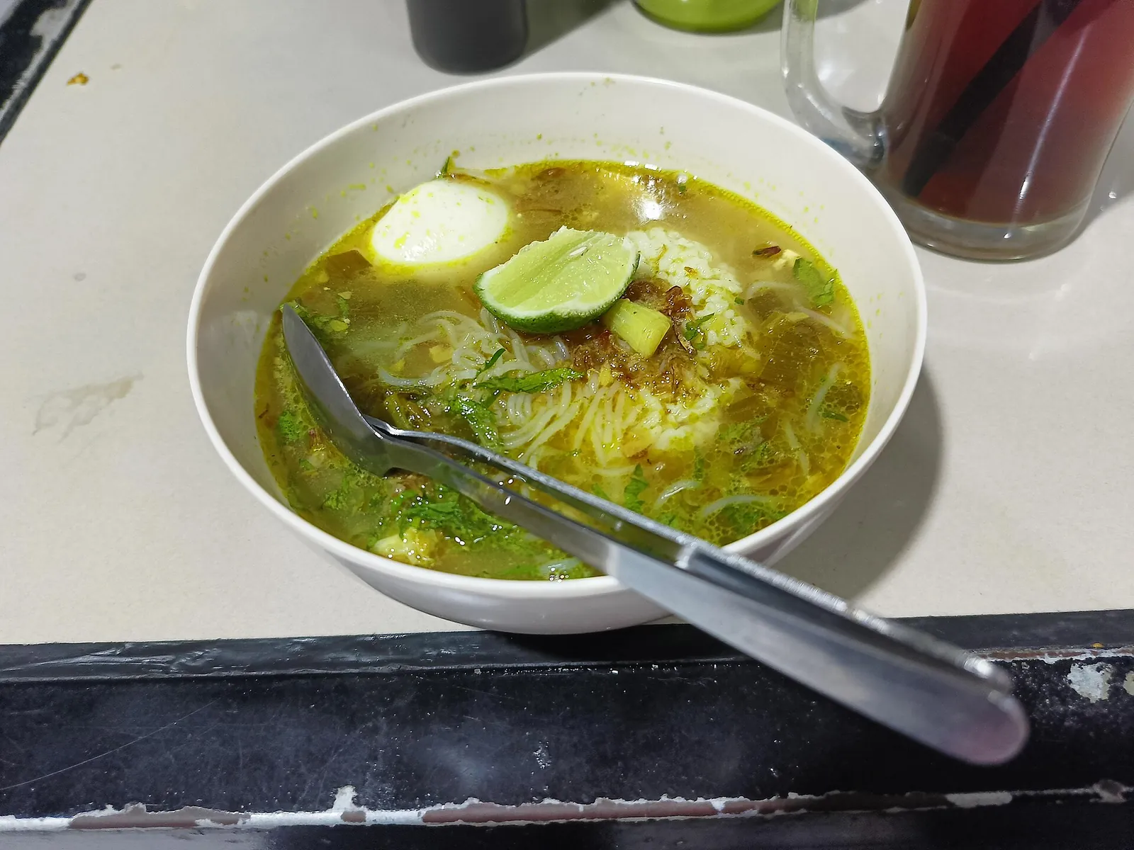A bowl of soto ayam with noodles, egg and a lime wedge, next to a glass of iced tea