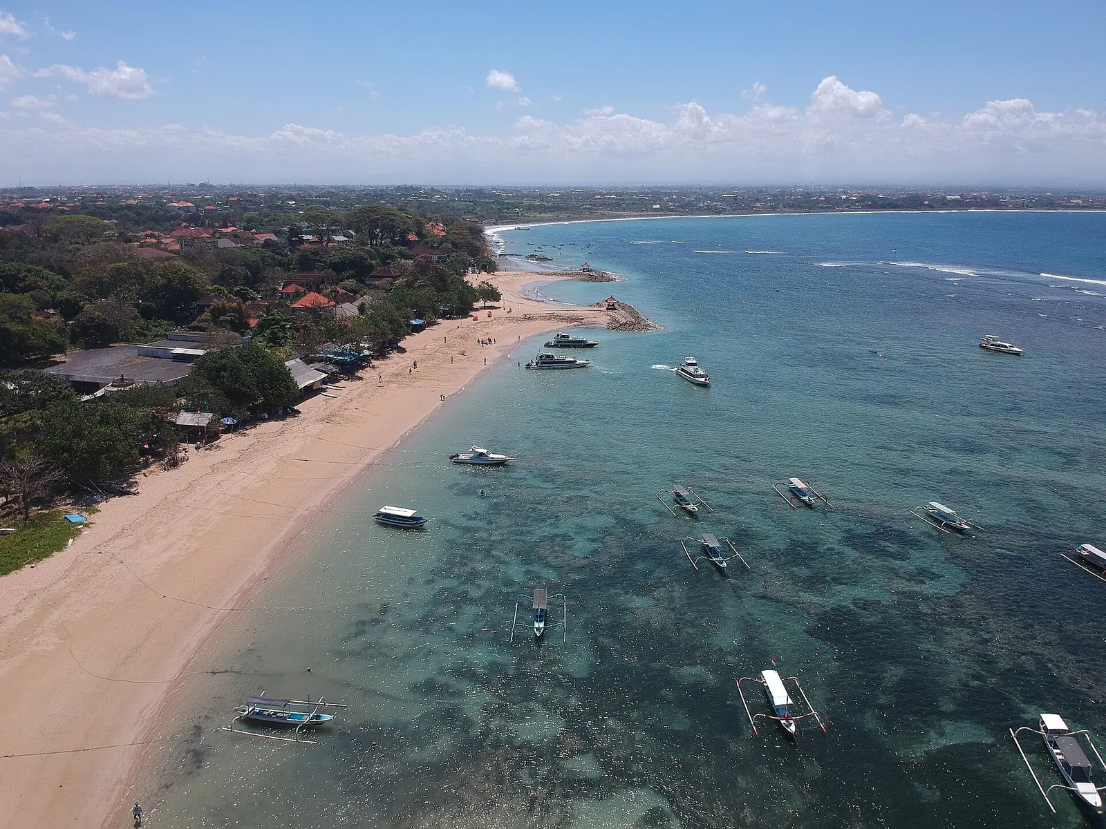 Aerial view of Sanur's coastline with a sandy beach, low-rise hotels among trees and boats moored over the reef lagoon