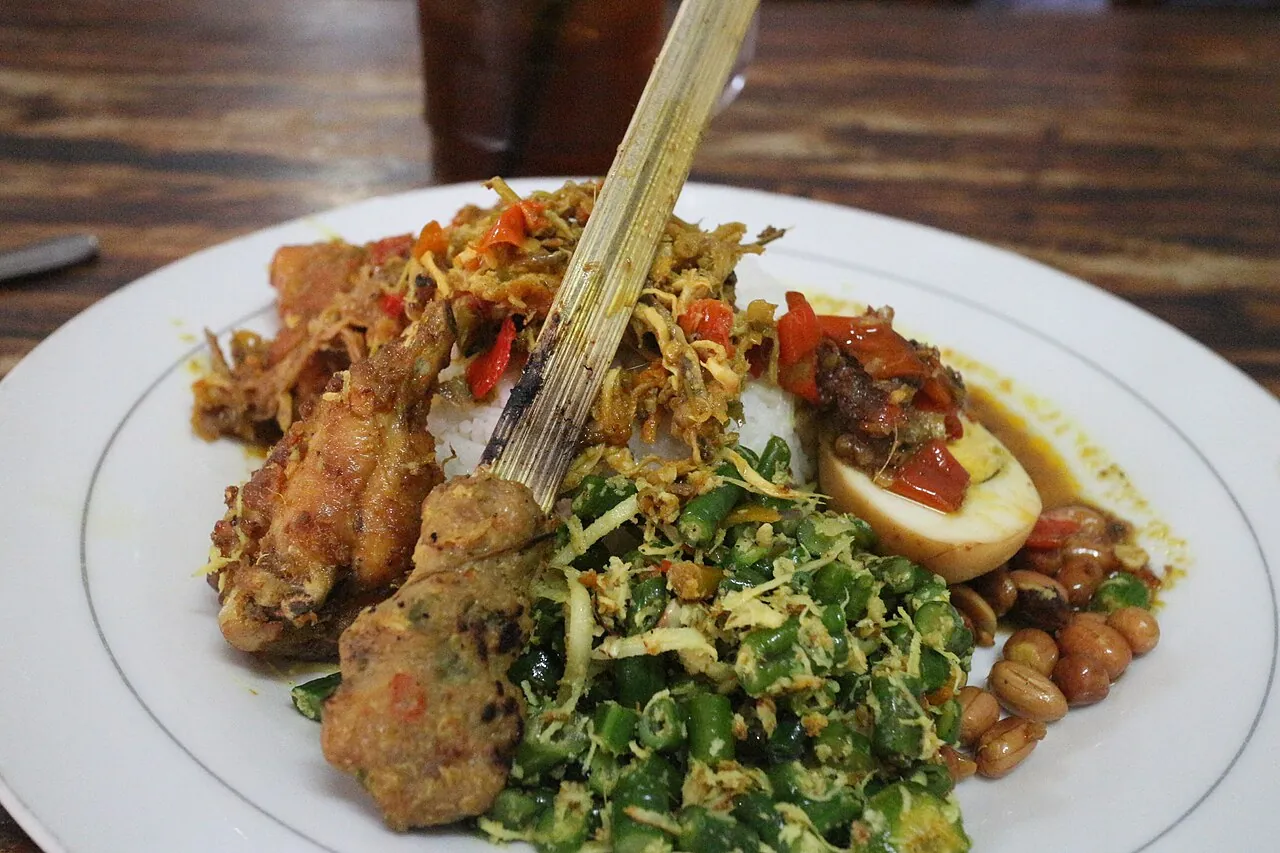 A plate of Balinese nasi campur with rice, fried chicken, sate lilit on a lemongrass stick, a spiced egg, green vegetables and peanuts