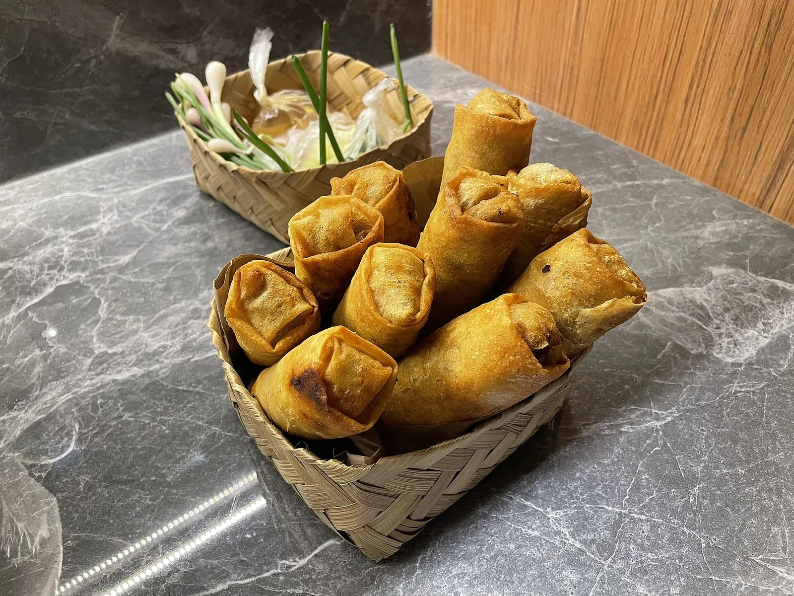 Fried Semarang lumpia spring rolls in a woven bamboo basket next to a basket of spring onions and sauce