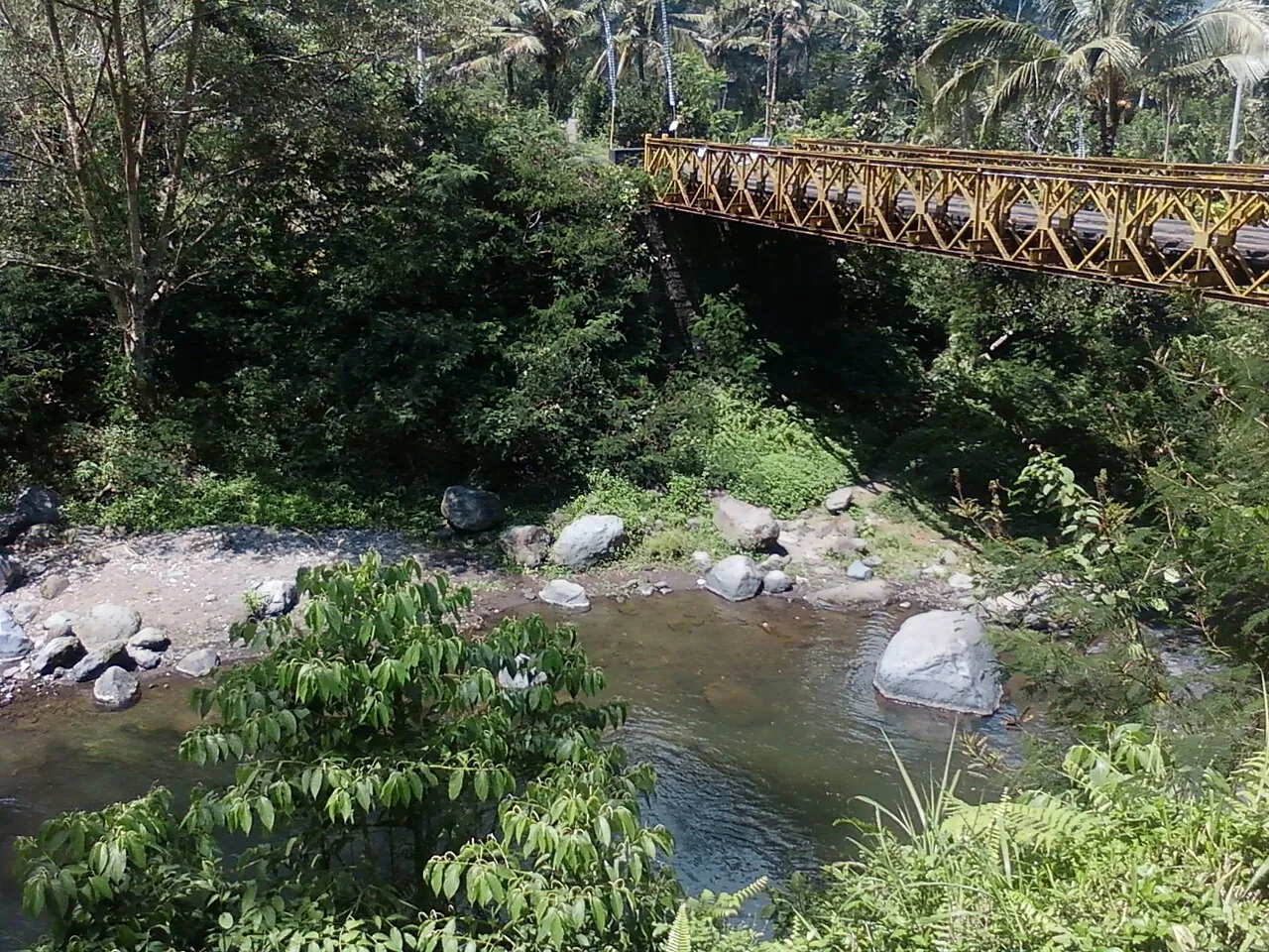 The rocky bed of the Tukad Unda river under a steel bridge, surrounded by green forest, at Sidemen
