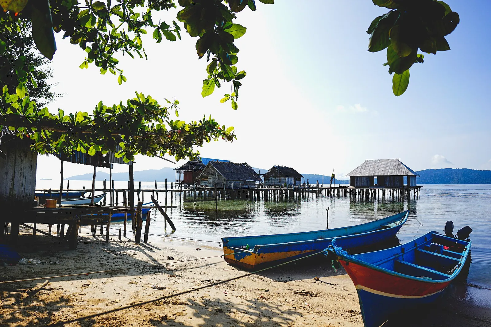 Wooden stilt homestays standing over clear blue water beside a beach with two moored boats in Sawinggrai, Raja Ampat