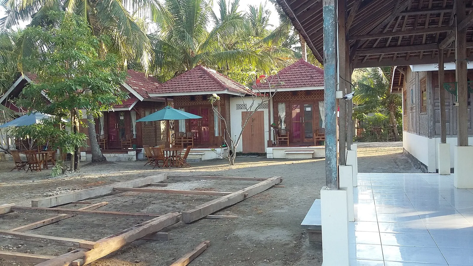 Small red-roofed homestay bungalows and a terrace with umbrellas among palm trees in Watukarung village, East Java