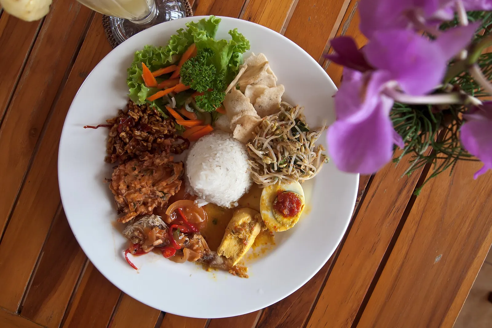 A plate of nasi campur with white rice, salad, bean sprouts, crackers, fried side dishes and a halved egg topped with chilli sauce, on a wooden table in Ubud