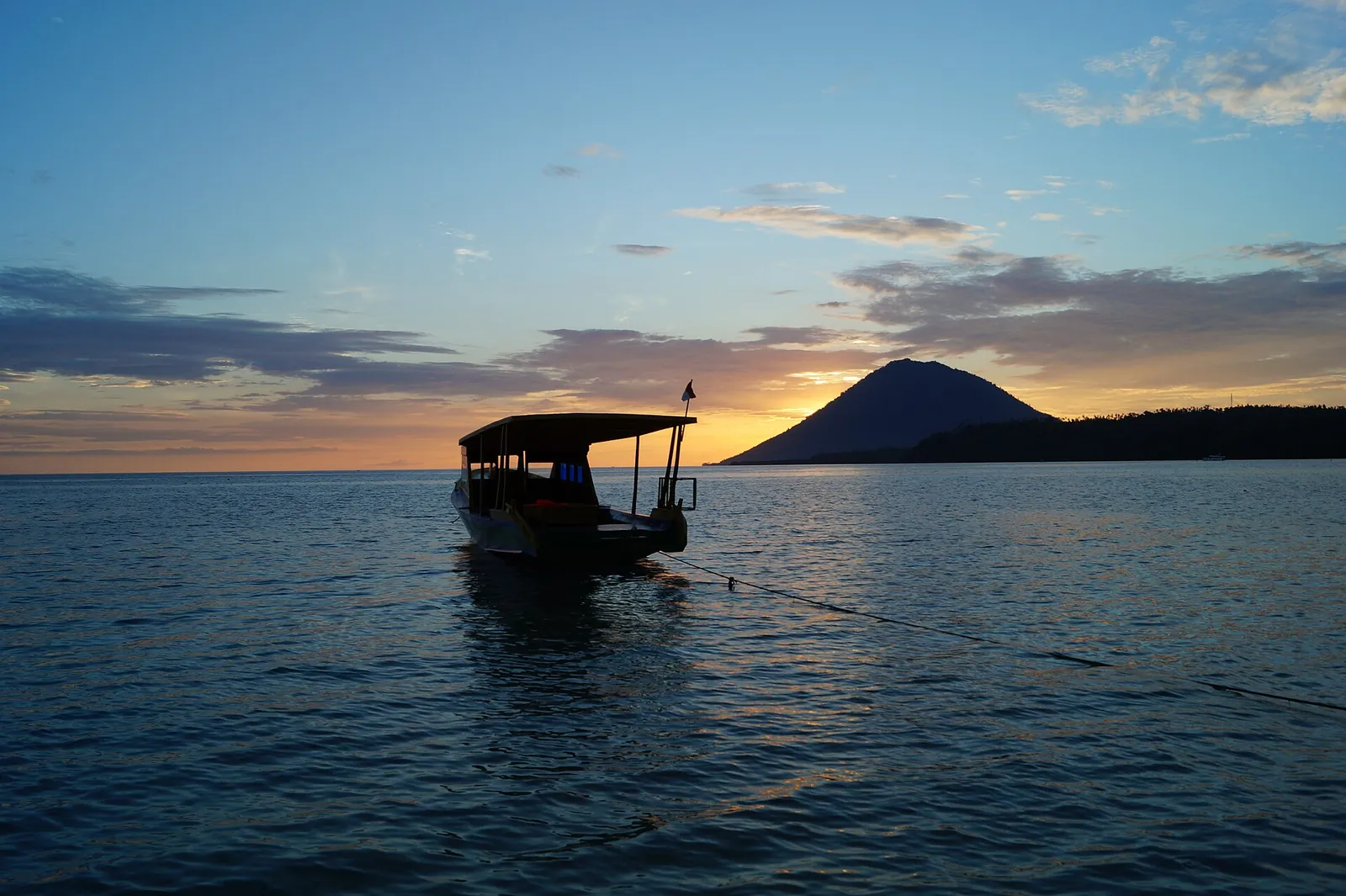 A small wooden boat on calm water at sunset off Bunaken, with the cone of Manado Tua on the horizon