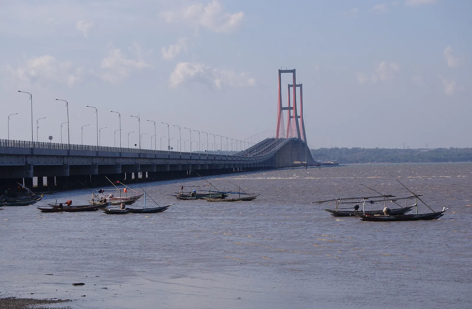 The Suramadu Bridge's red twin towers over the Madura Strait, with wooden fishing boats in the foreground