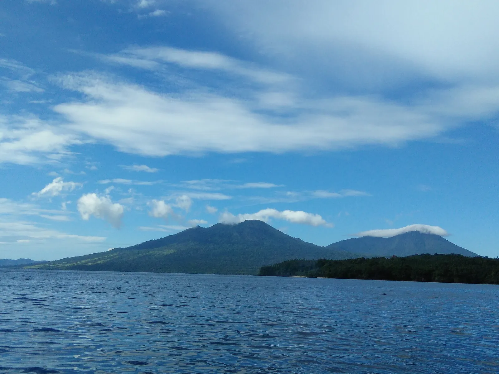 Two forested volcanic peaks of Tangkoko and Duasaudara seen across the sea under blue sky