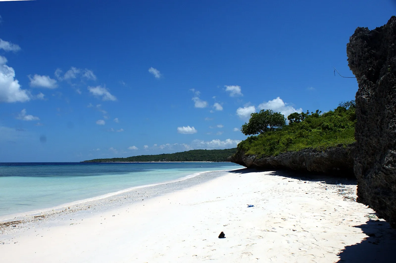 A white-sand beach below a wooded limestone headland at Bira under a blue sky