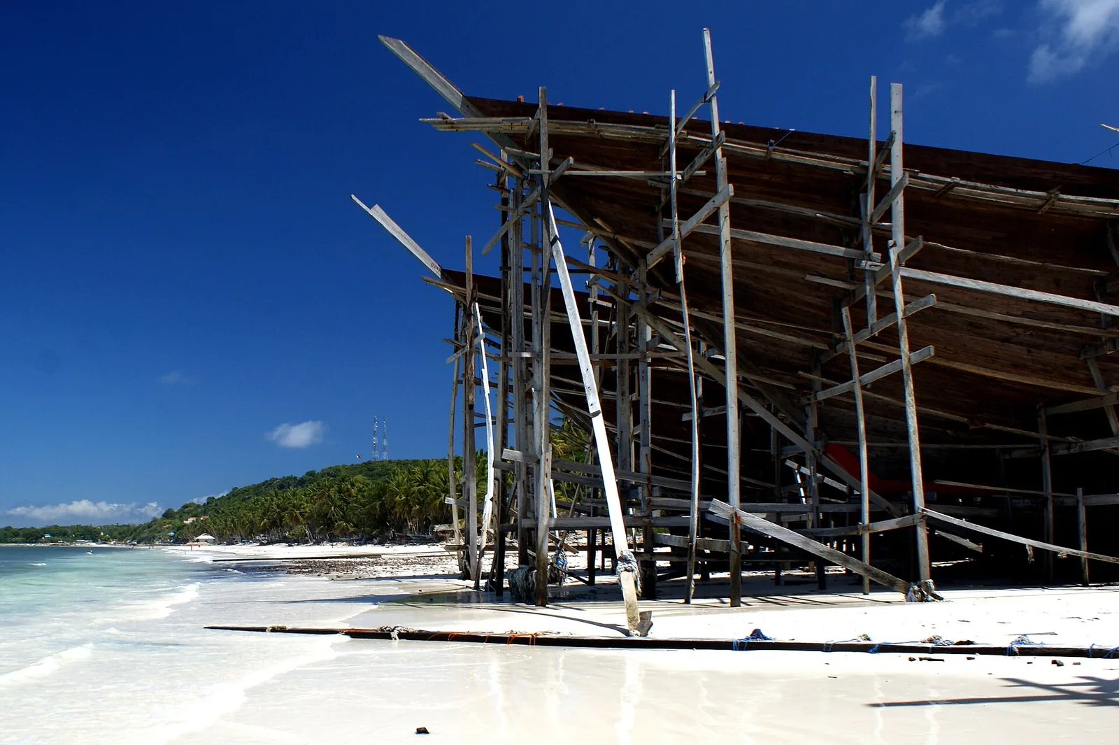 A wooden hull under construction on a timber scaffold beside a white beach at Bira