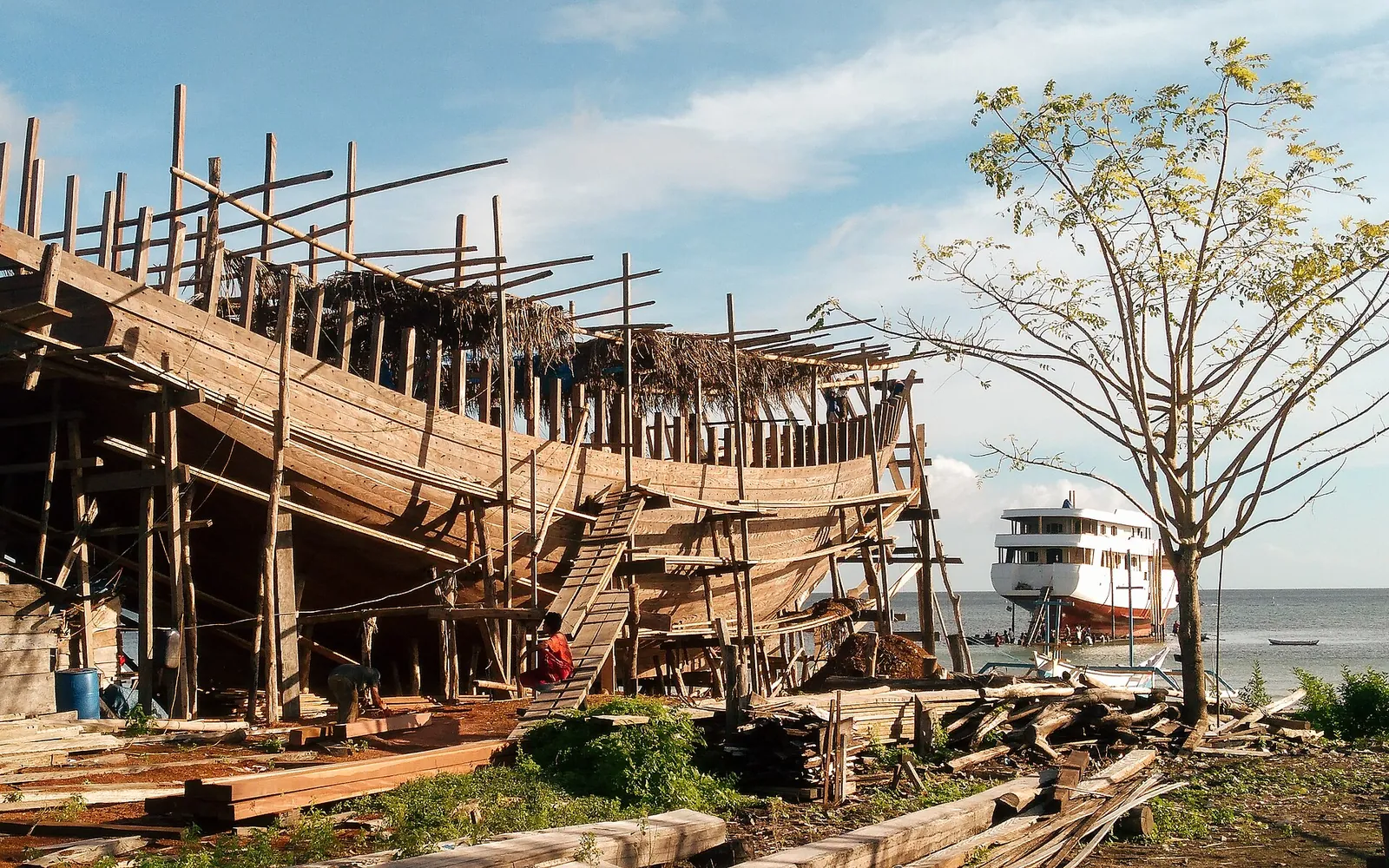 The wooden hull of a pinisi on its building frame on the shore at Tanaberu, with a finished boat on the water behind