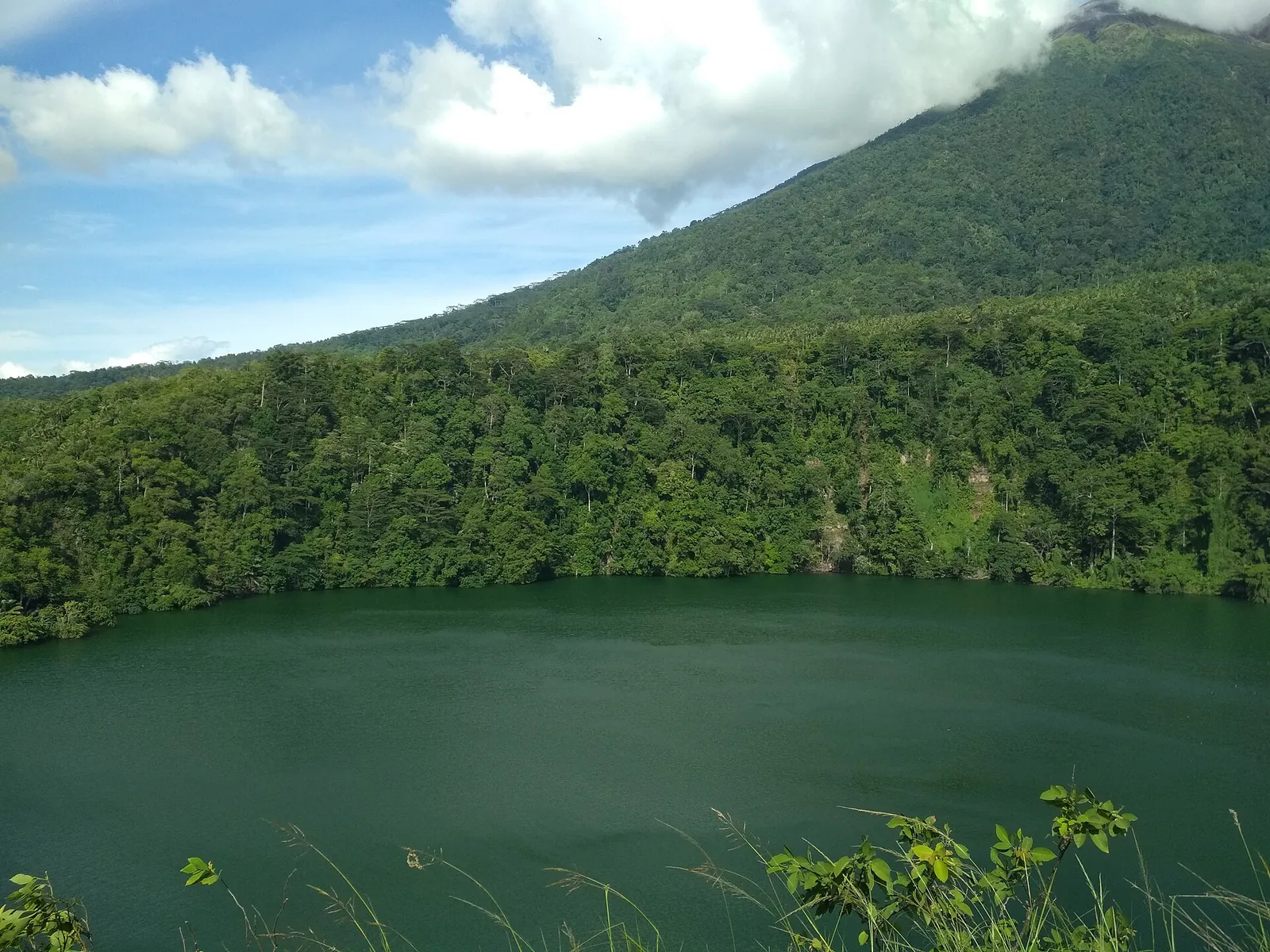 The green crater lake of Tolire Besar ringed by forest, with the slopes of Mount Gamalama behind