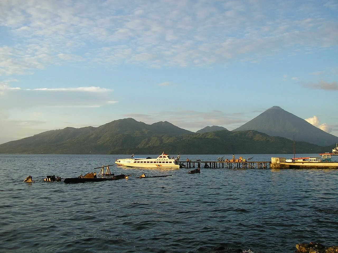 Tidore's volcanic peak across the strait from Ternate, with a passenger boat at a jetty in the foreground