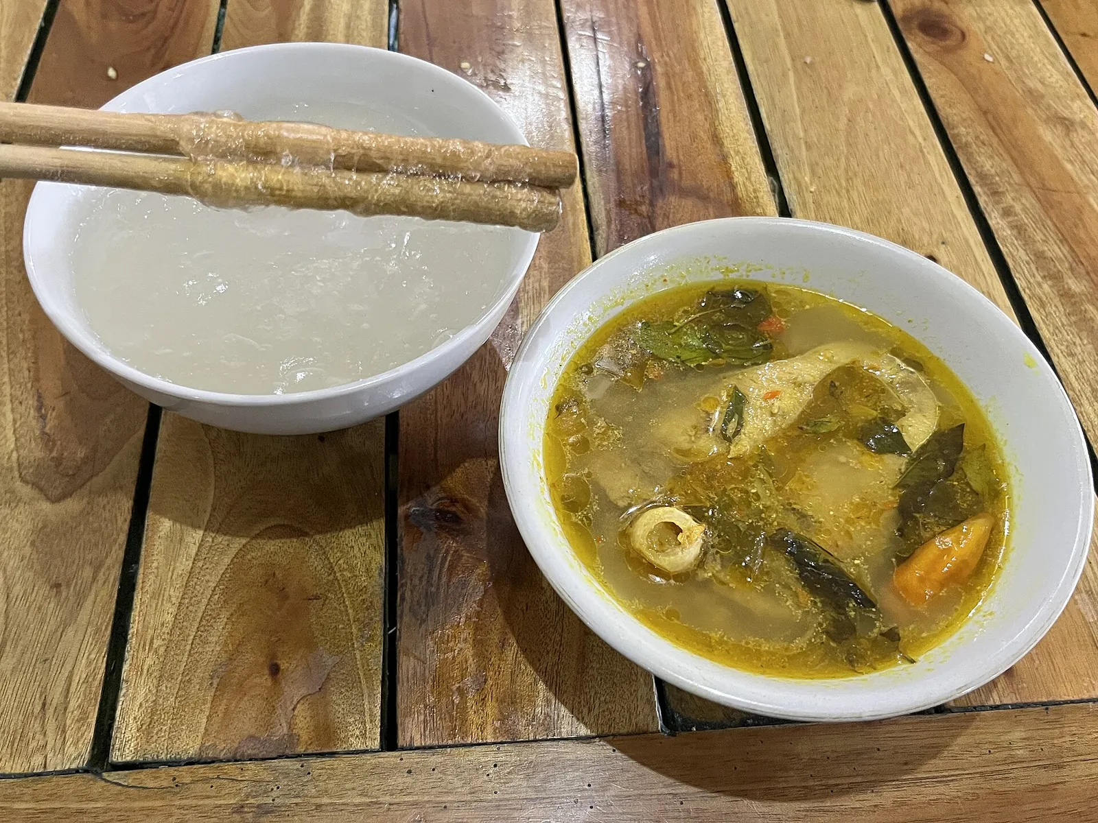 A bowl of clear, glossy papeda with bamboo chopsticks beside a bowl of fish in yellow turmeric broth