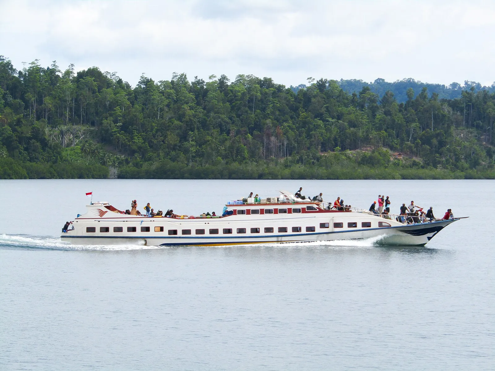 White passenger fast boat KM Mbura-Mbura crossing calm water in front of a forested shore