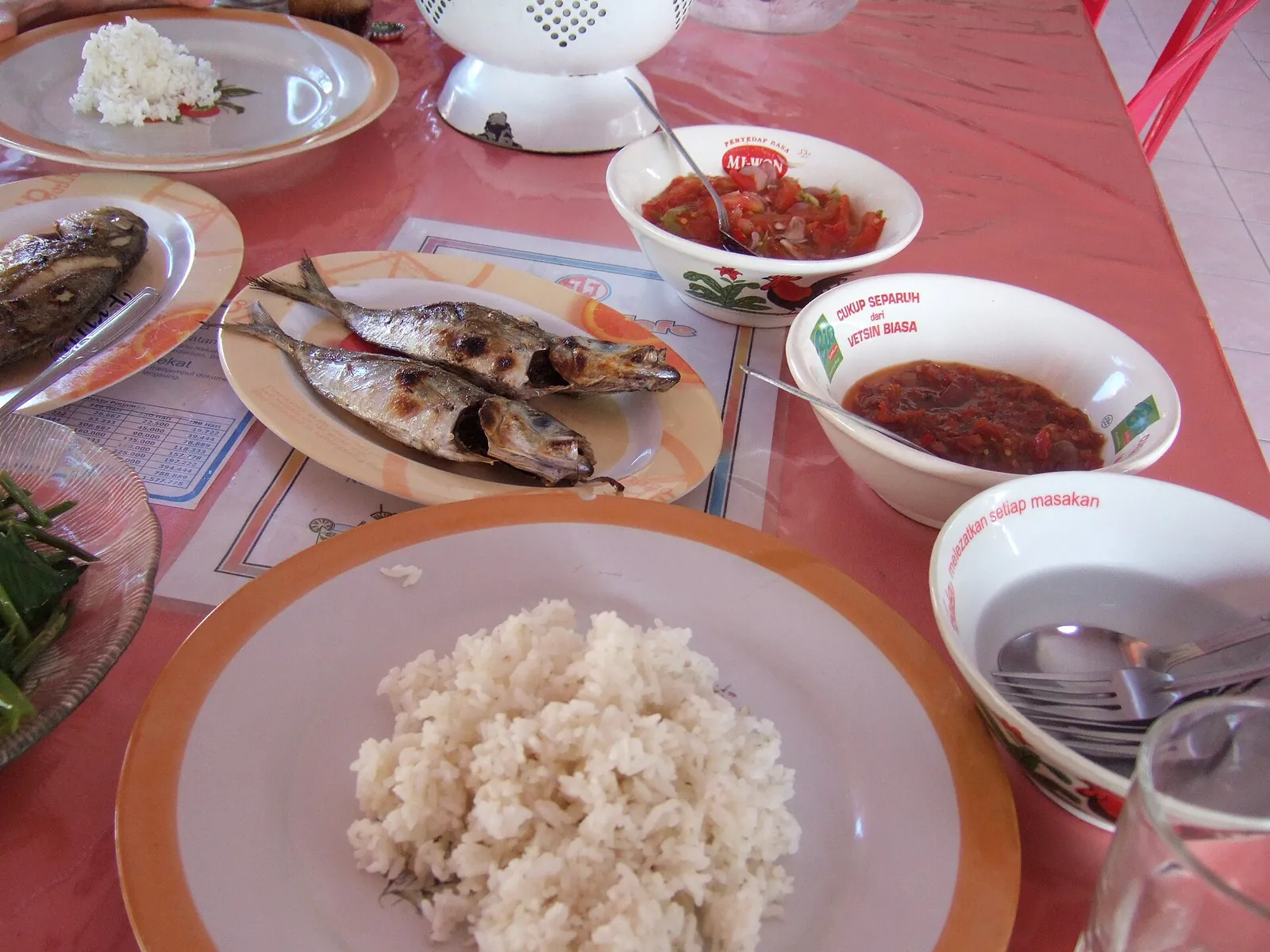 Plates of grilled fish with steamed rice and bowls of red chilli sambal on a table