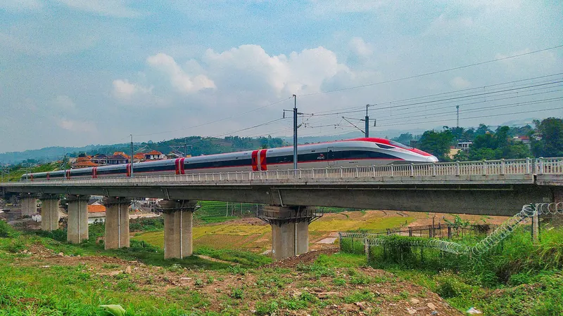 Red-and-white Whoosh high-speed train crossing a concrete viaduct over green fields in West Bandung Regency