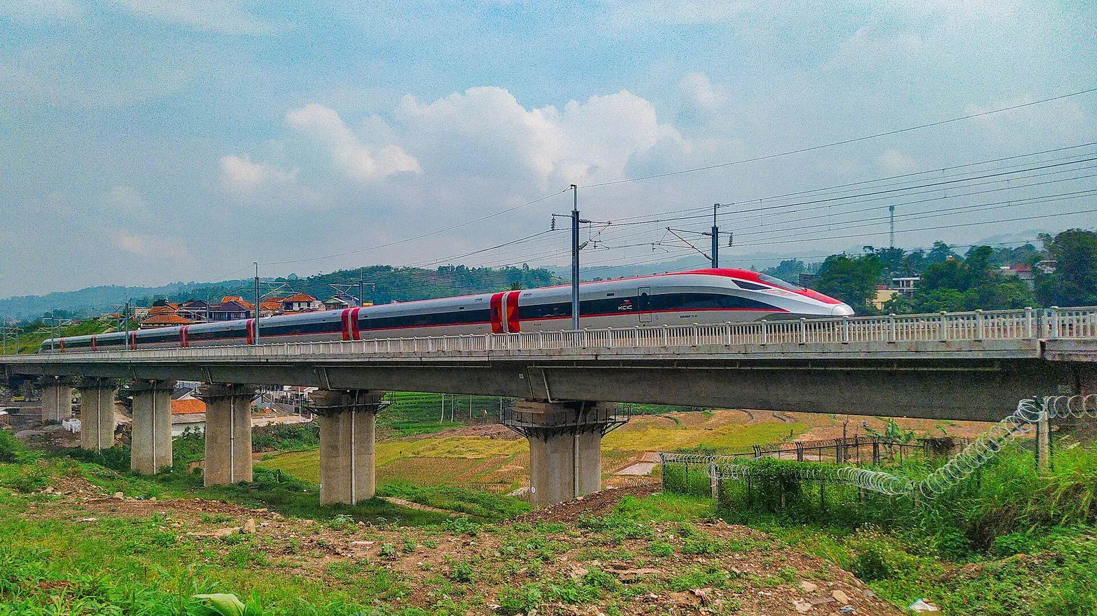 Red-and-white Whoosh high-speed train crossing a concrete viaduct over green fields in West Bandung Regency