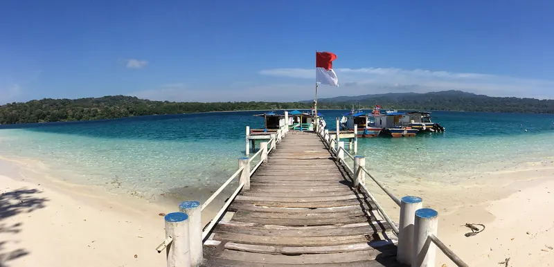 Wooden jetty with a red-and-white flag running out over turquoise water from the white-sand beach of Peucang Island