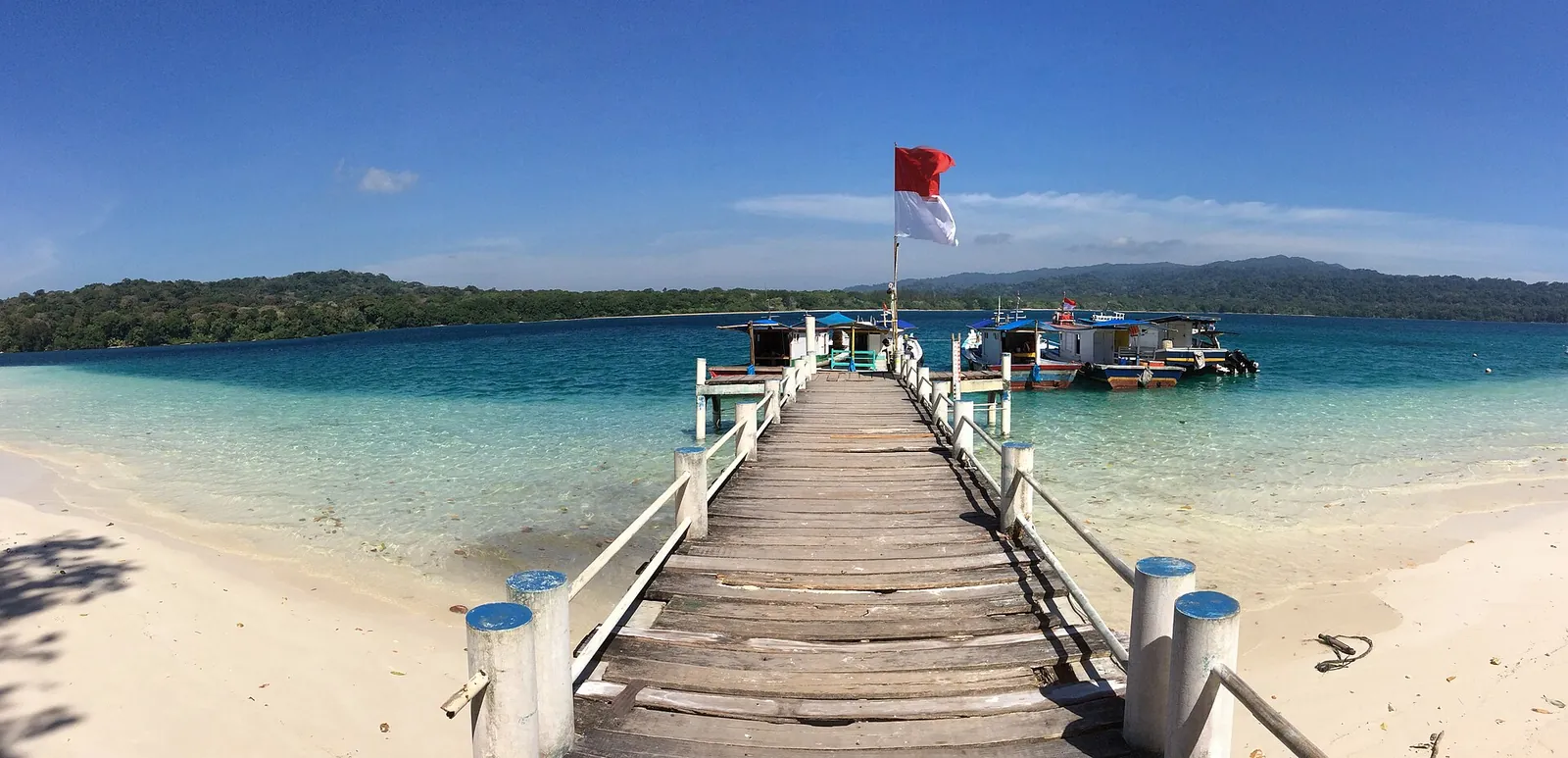 Wooden jetty with a red-and-white flag running out over turquoise water from the white-sand beach of Peucang Island