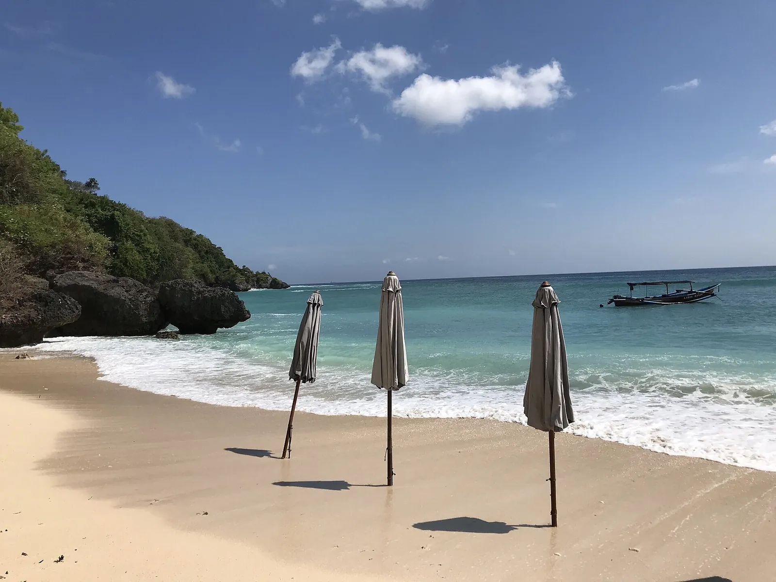 Beach umbrellas on the white sand of Padang Padang Beach with a wooded headland and a boat offshore