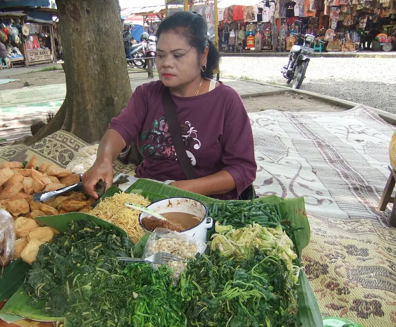 A pecel vendor sitting behind trays of boiled greens, bean sprouts and fried snacks at the Borobudur temple complex