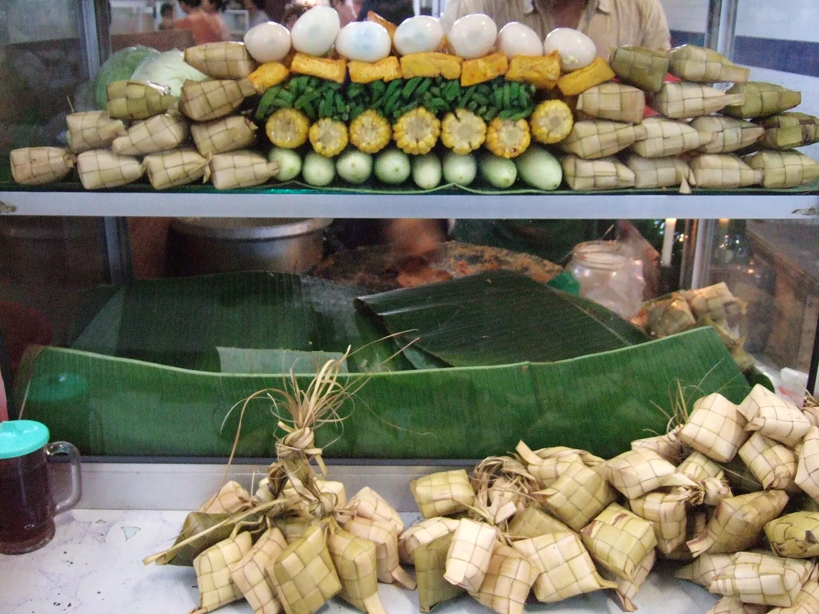 A gado-gado stall at a Jakarta market with ketupat rice cakes hanging, and potatoes, eggs and vegetables stacked behind glass