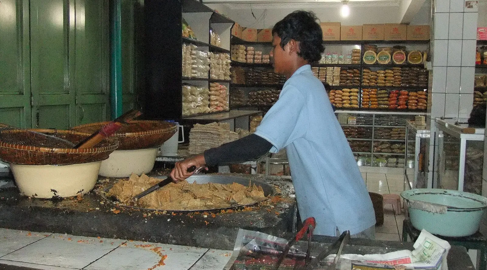 A cook frying tempeh chips in a large wok in a shop in Lembang, with packed snacks on shelves behind