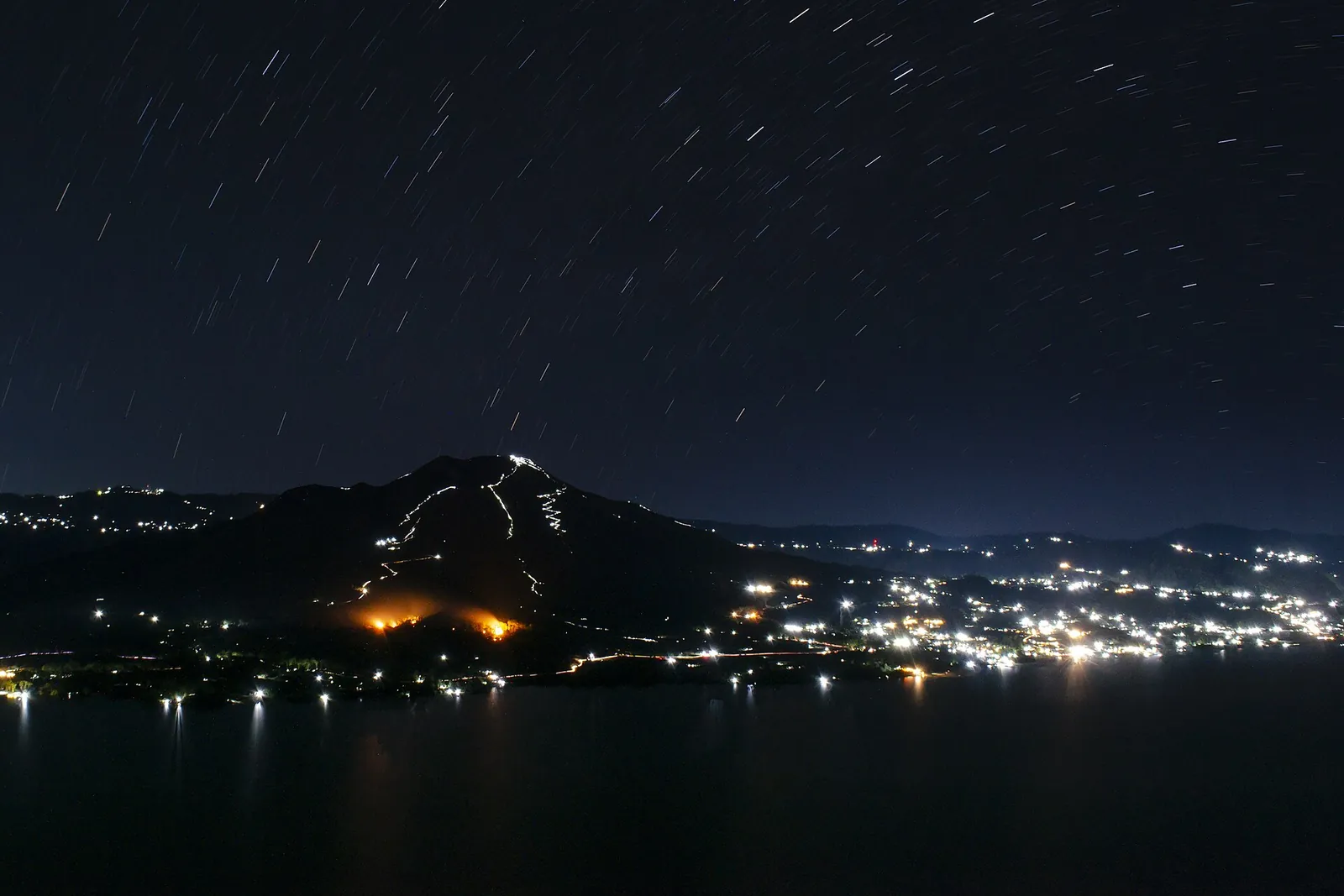 Star trails over Mount Batur at night, with a line of hikers' torches on the slope and village lights below