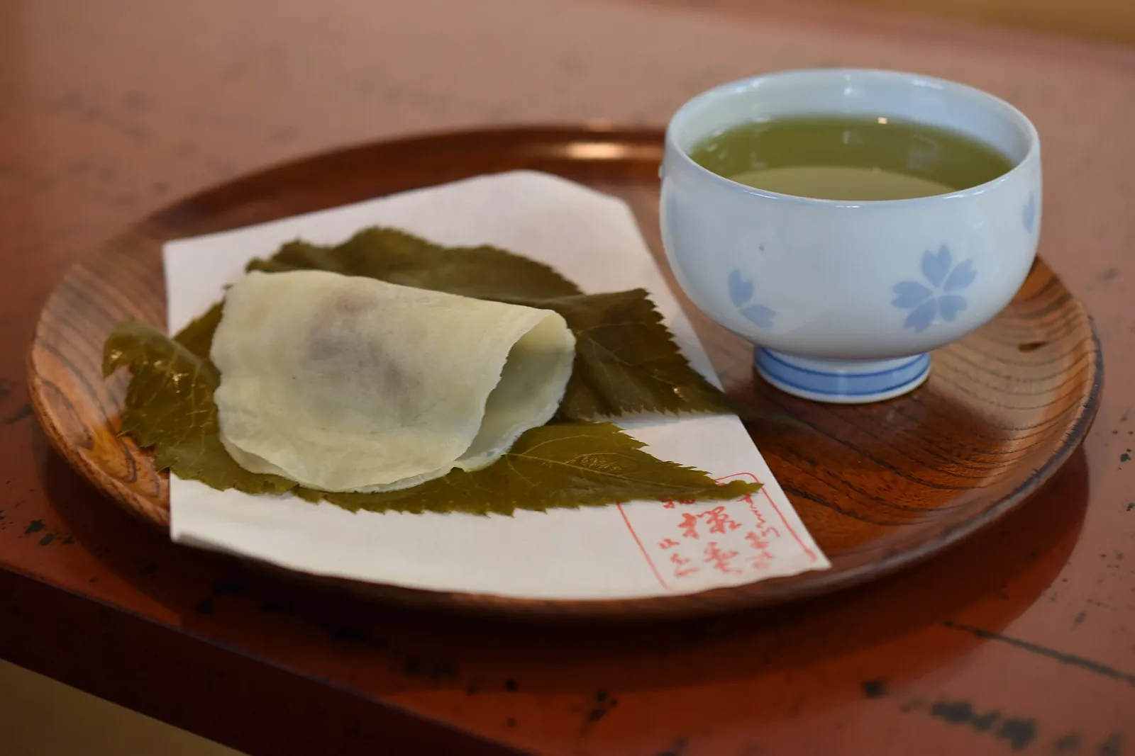 Sakura mochi ve stylu Chomeiji zabalené v třešňových listech na dřevěném podnose vedle šálku zeleného čaje