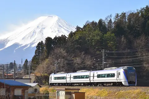Bílo-fialový expres Fuji Excursion jedoucí kolem borových lesů a domů, za ním se pod jasně modrou oblohou zvedá zasněžená hora Fudži