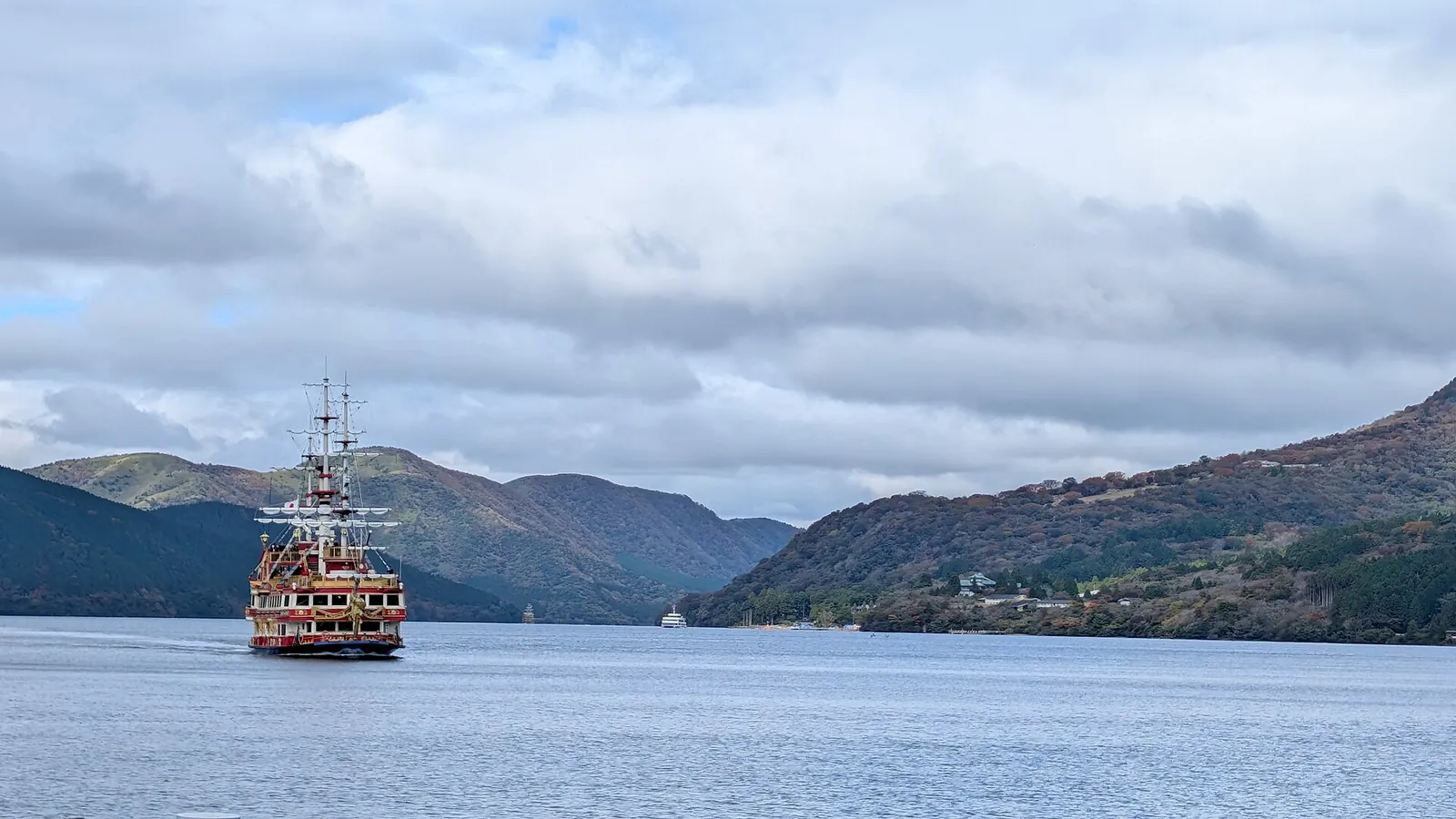 Červeno-zlatá vyhlídková loď ve stylu pirátské lodi křižující jezero Ashi v Hakone, za ní podzimně zbarvené kopce a zataženou oblohu