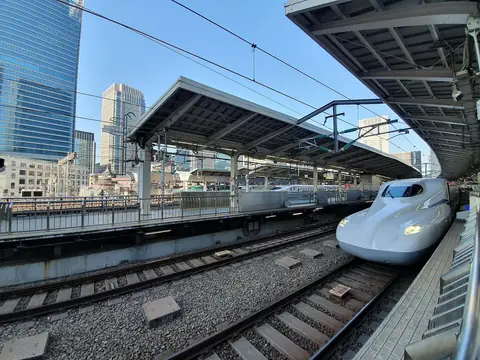 Bílý shinkansen vjíždějící na otevřené nástupiště Tokijského nádraží, s kancelářskými věžemi vzadu pod jasně modrou oblohou