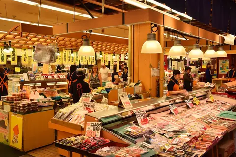 Un mostrador de marisco en el Mercado Omicho de Kanazawa bajo lámparas colgantes, con bandejas de pescado y huevas con precio en la vitrina refrigerada y personal con delantales naranjas atendiendo a los clientes