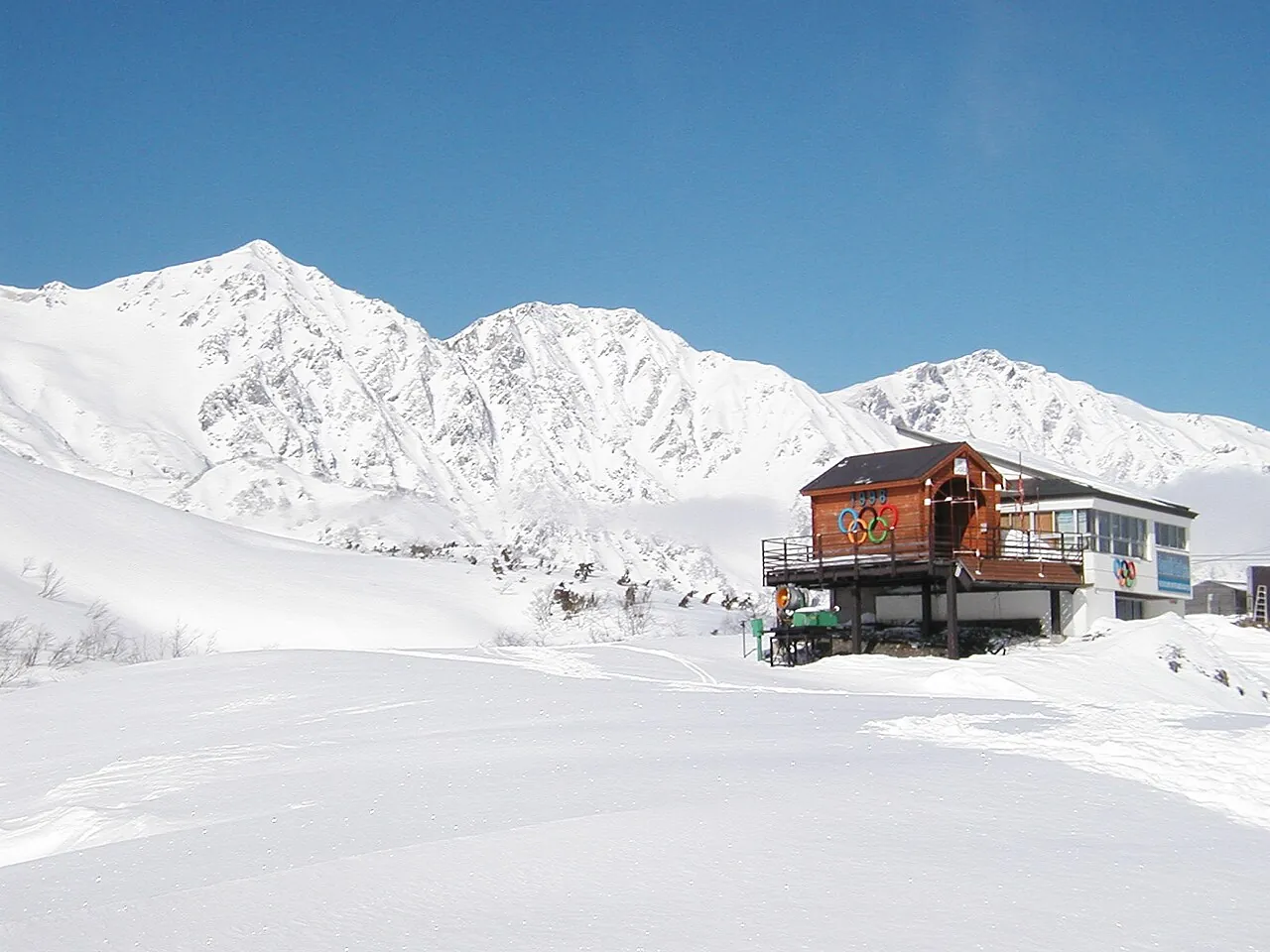 Un abri de départ en bois orné des anneaux olympiques sur une pente enneigée de Happo-one, avec les pics blancs de Hakuba derrière sous un ciel bleu