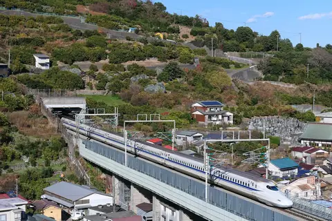 Bílý shinkansen N700A na viaduktu mezi Odawarou a Atami, projíždějící kolem domů a svahových sadů pod modrou oblohou