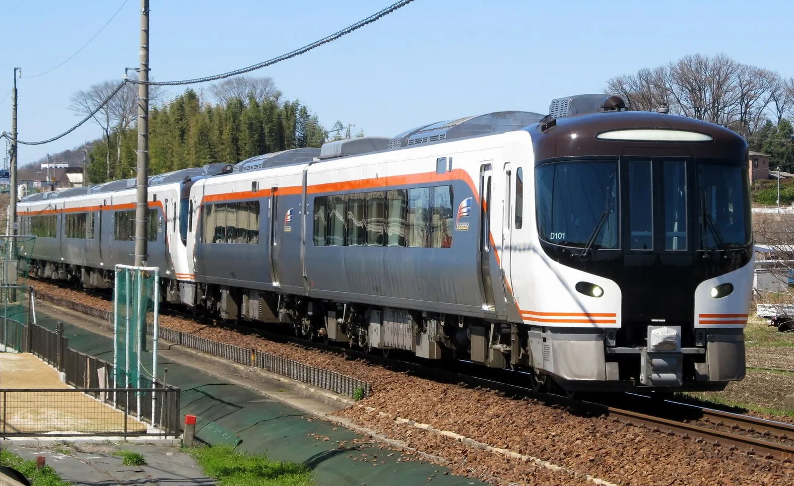 Bílo-oranžový expres HC85 Hida projíždějící malou stanicí na trati Takayama pod modrou oblohou