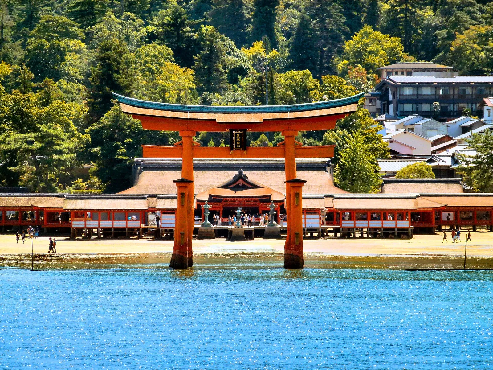 The red torii of Itsukushima Shrine standing in blue water in front of the shrine and green hills