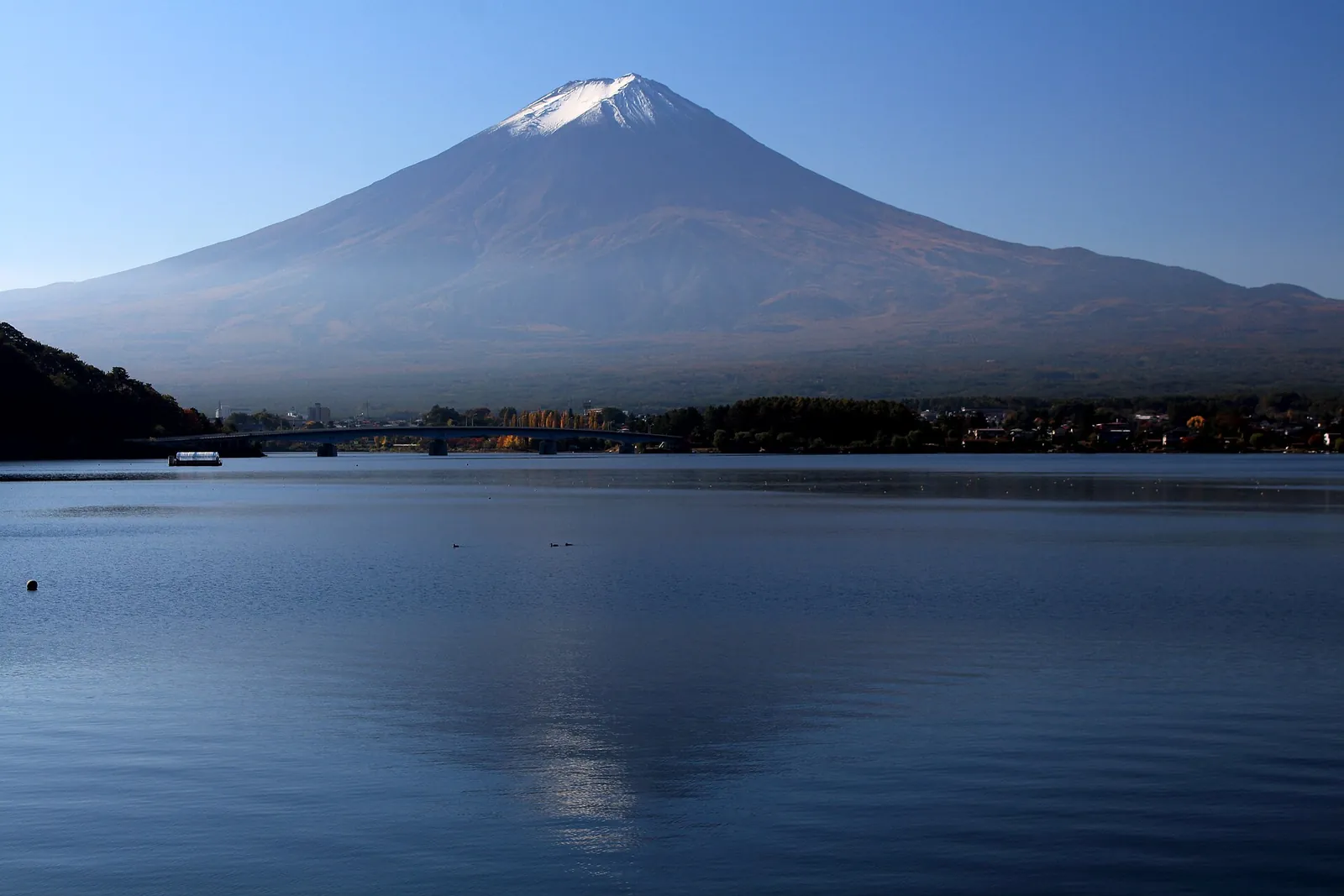 Zasněžená hora Fuji tyčící se nad klidnou hladinou jezera Kawaguchi pod jasnou modrou oblohou, s zalesněným vzdáleným břehem dole