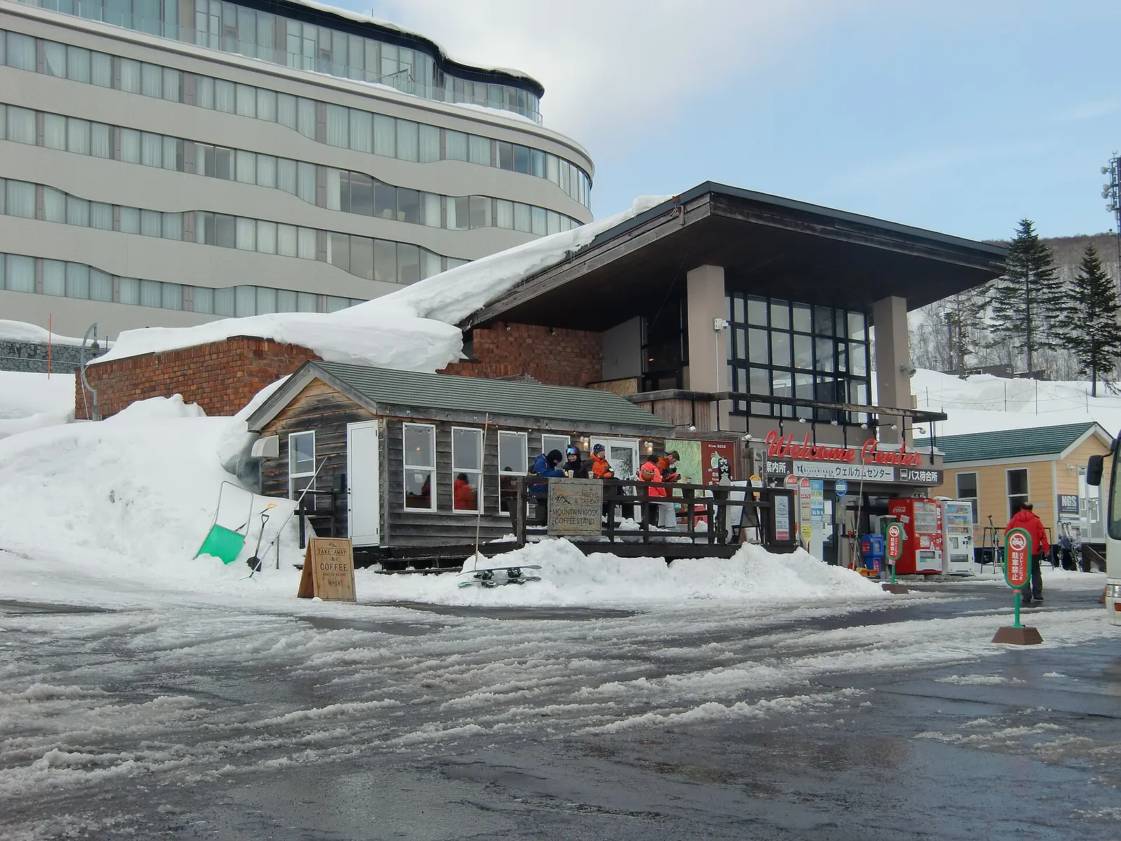 Grand Hirafu Welcome Center, nízká budova s obchodem a autobusovou zastávkou, před velkým hotelem s navátým sněhem kolem