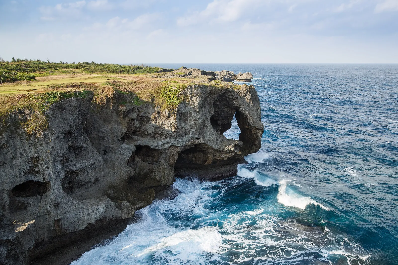 Acantilados de piedra caliza y una roca con arco en el cabo Manzamo, en la costa de Onna, con la blanca espuma rompiendo sobre el agua de un azul intenso
