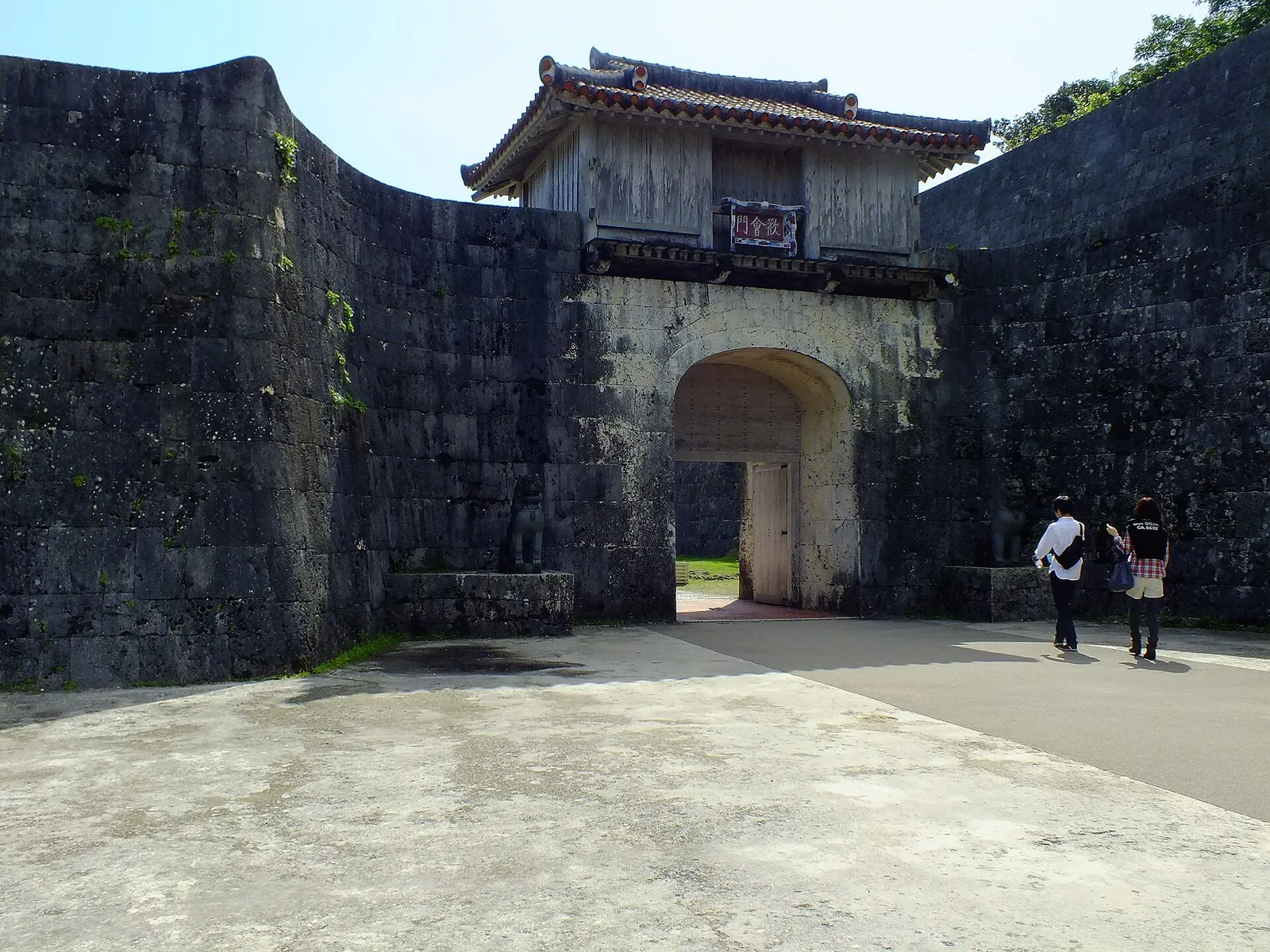 La puerta Kankaimon, de muros de piedra, del castillo de Shuri: una caseta de entrada de madera sobre una abertura en arco en un muro de piedra caliza gris, bajo un cielo azul