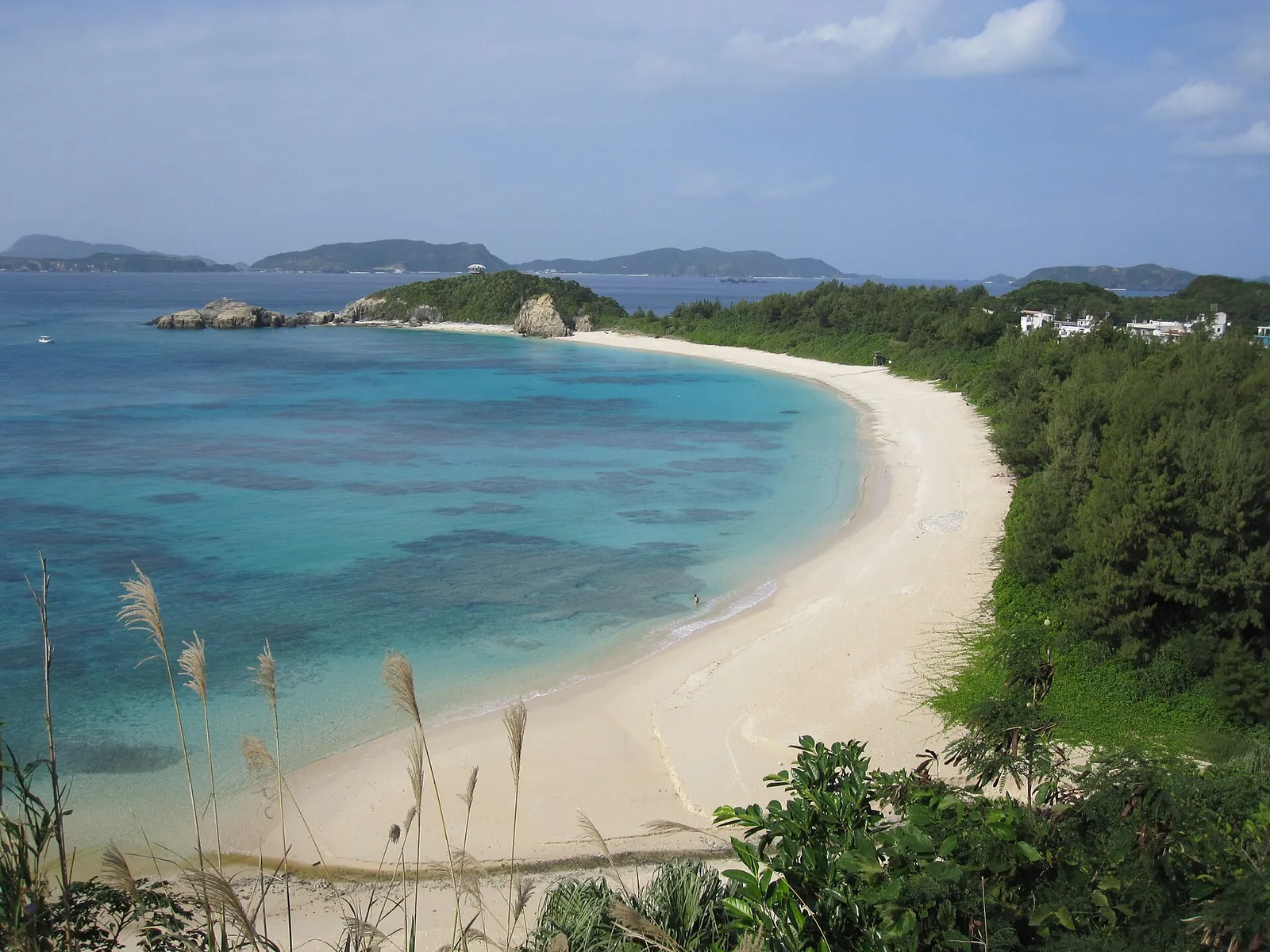 La playa de Aharen en la isla de Tokashiki: una curva de arena clara junto a aguas turquesas, con colinas boscosas e islotes al fondo