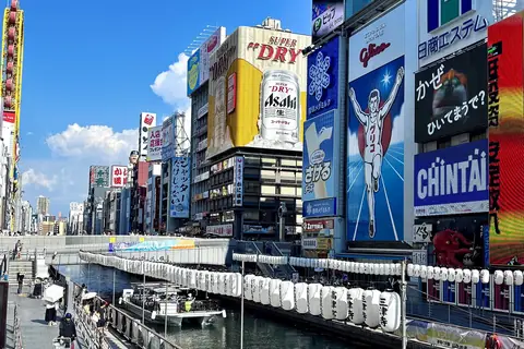 Der Kanal von Dotonbori in Osaka unter blauem Himmel, gesäumt von riesigen Werbetafeln, darunter der Glico-Läufer, mit einem Uferweg und einer Reihe weißer Laternen