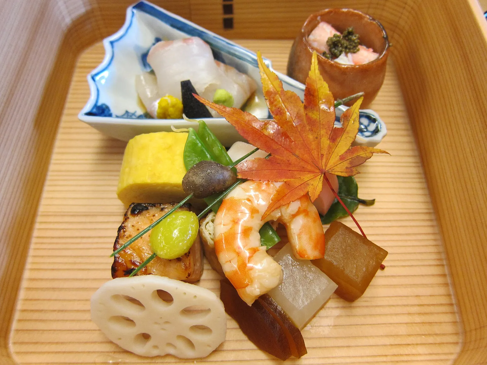 Chod kaiseki z malých sezonních jídel v dřevěné krabičce: sašimi, rolovaná omeleta, kreveta, grilovaná ryba, kořen lotosu a dušená zelenina pod červeným javorovým listem