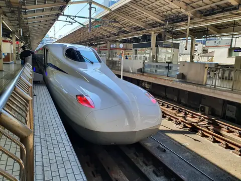 Bílý shinkansen N700S stojící u nástupiště 18 na nádraží Tokio, jeho dlouhý špičatý nos vedle zábradlí nástupiště pod stříškou nádraží